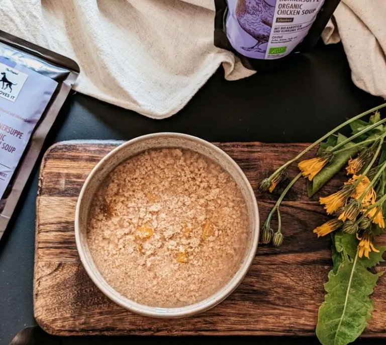 Organic chicken soup in a bowl on a wooden board, surrounded by flowers and packaging.